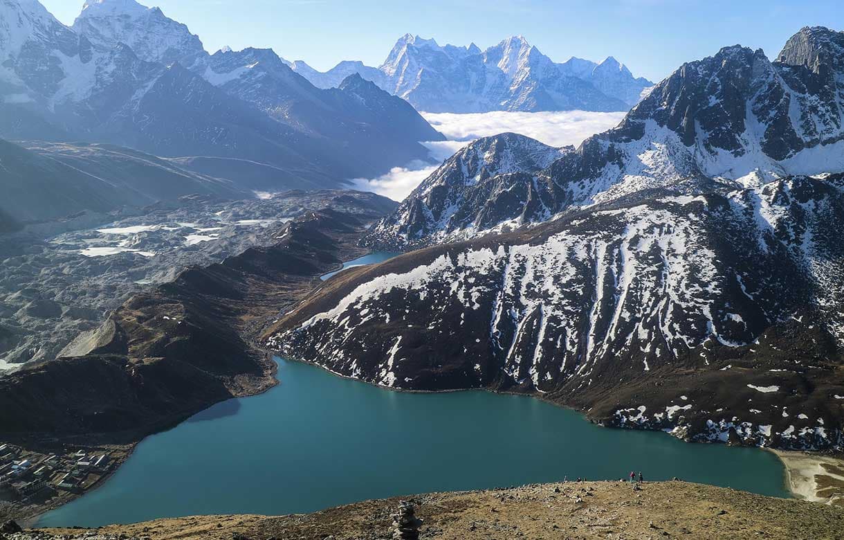 Panoramic high-altitude view of a turquoise glacial lake next to a massive glacier and snow-capped Himalayan mountains.