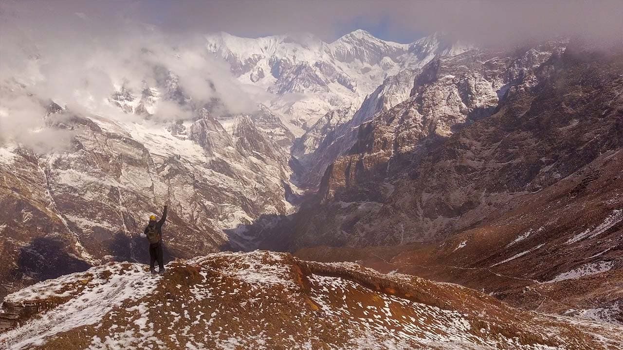 A trekker standing on a snowy ridge with raised arm, looking at dramatic snow-capped mountains and valleys.
