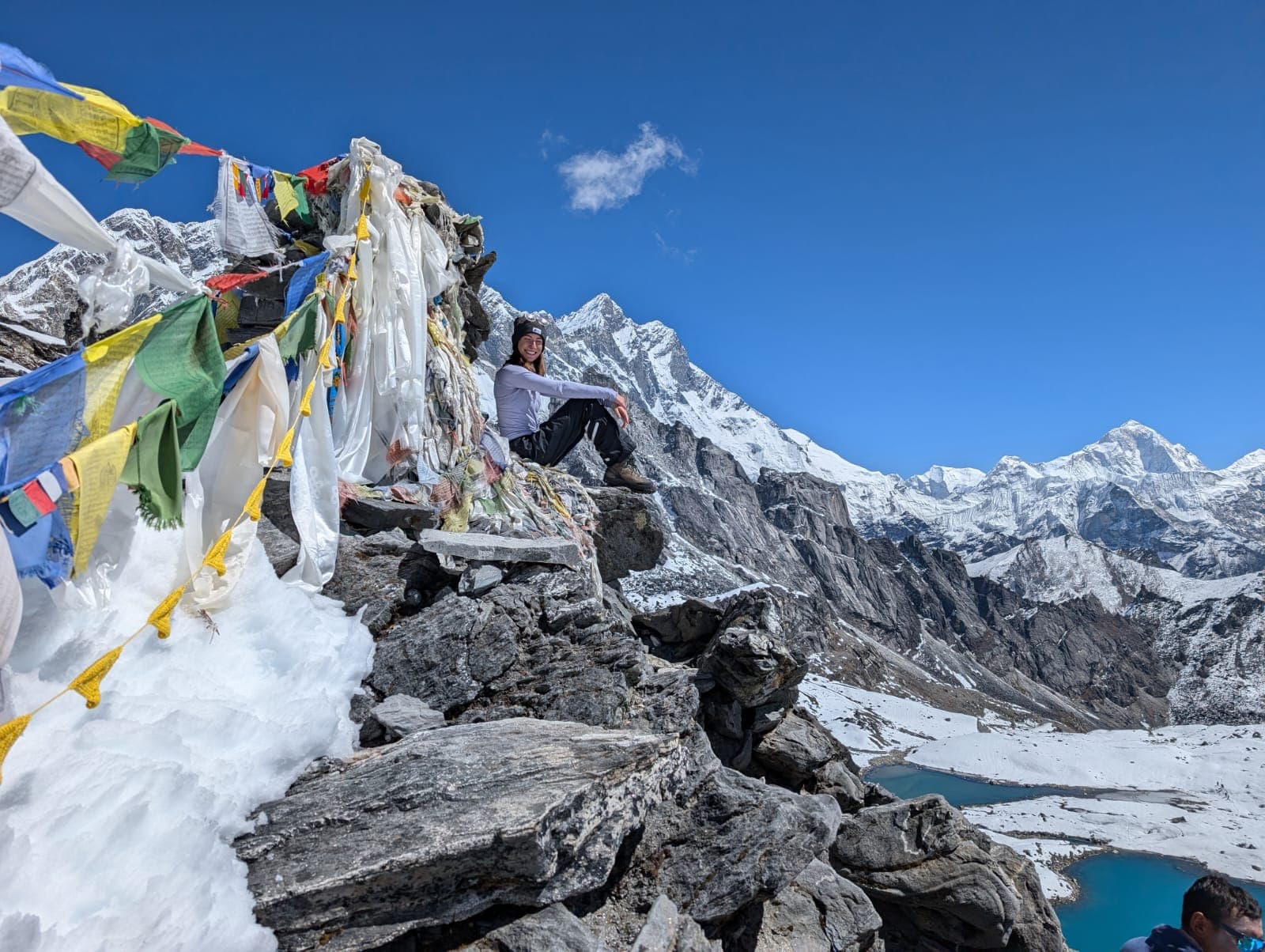 A female trekker sitting on a rocky peak beside prayer flags, overlooking snow-covered Himalayan peaks and blue lakes.