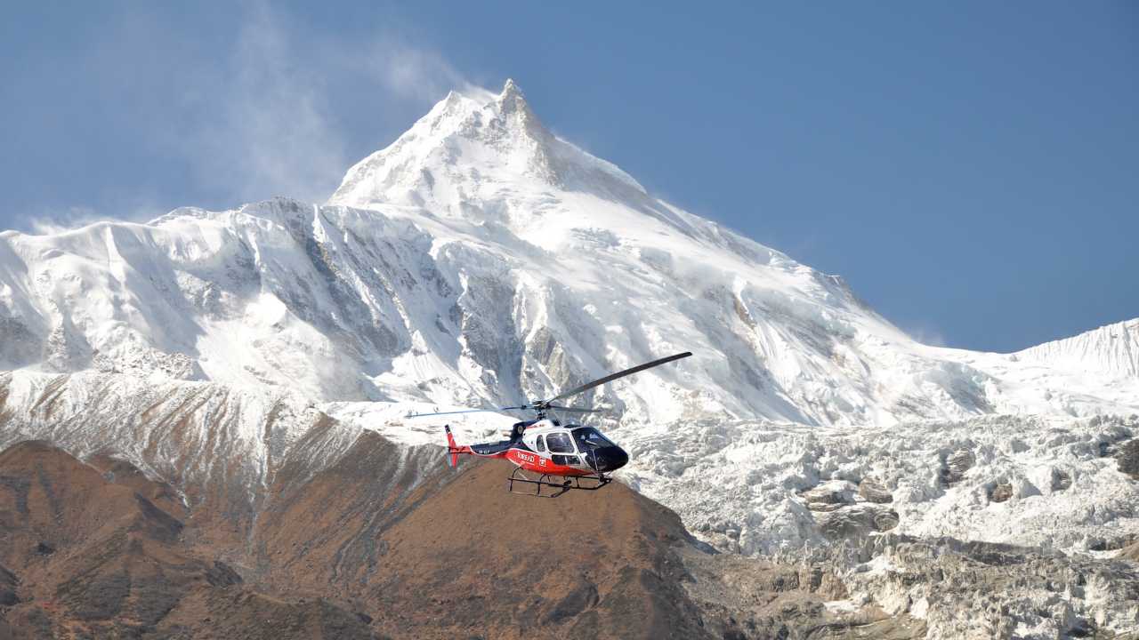 Red and white helicopter flying past a snow-capped Himalayan peak in the Manaslu region.