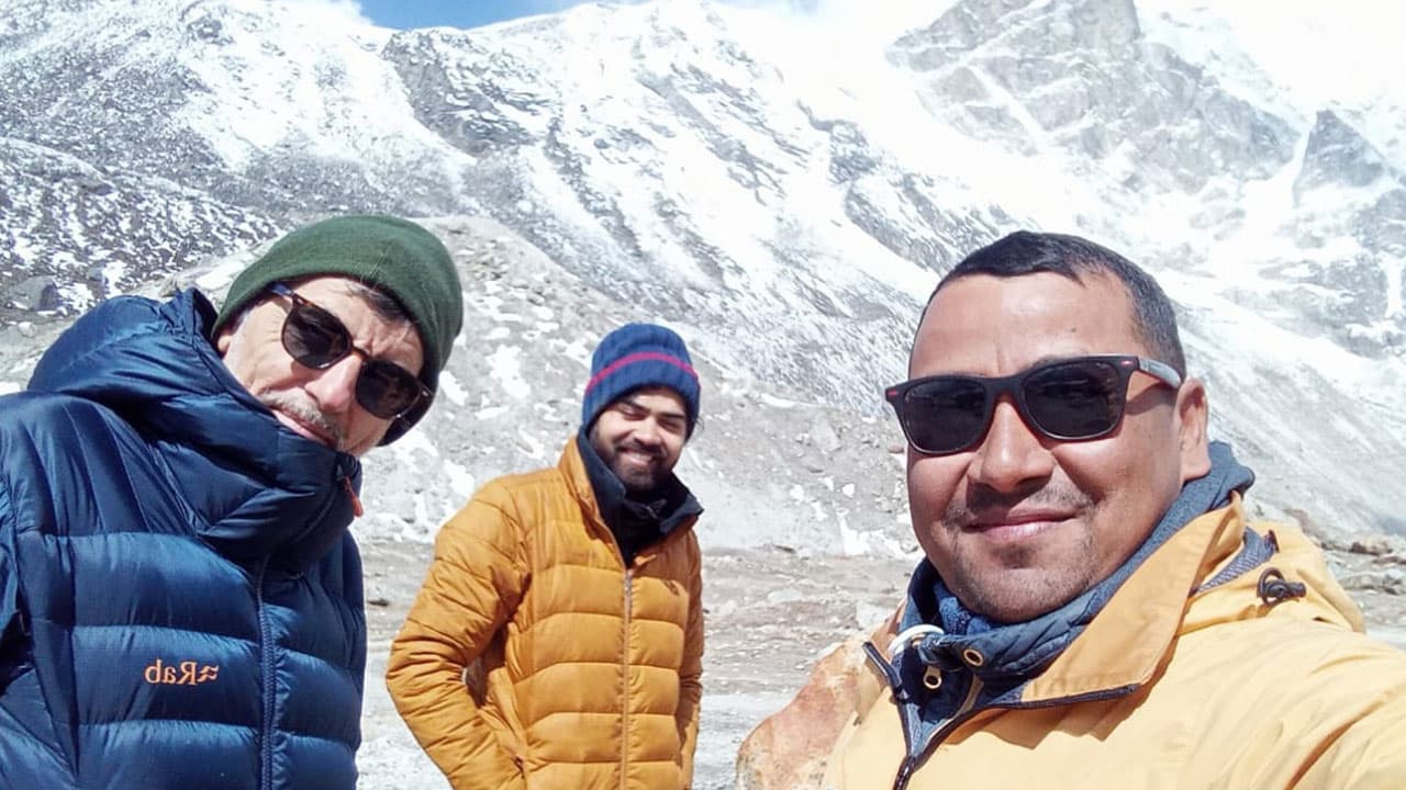 Trekkers smiling for a selfie wearing winter gear and sunglasses in front of snowy Himalayan peaks during the Makalu High Pass Trek.
