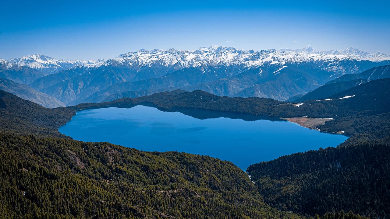 High-altitude aerial view of Rara Lake surrounded by green forest with snow-capped mountains in the distance.