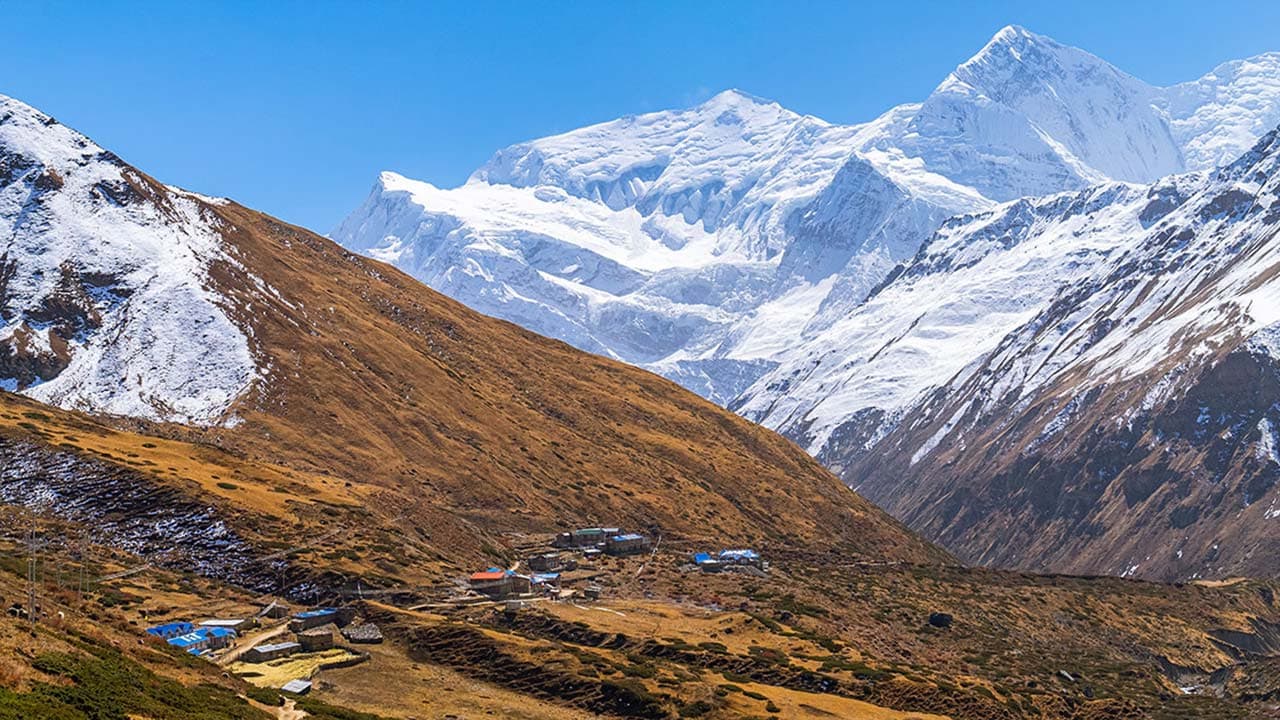 Small high-altitude Himalayan village on brown hillsides with snow-capped Annapurna mountains in the background under a blue sky.