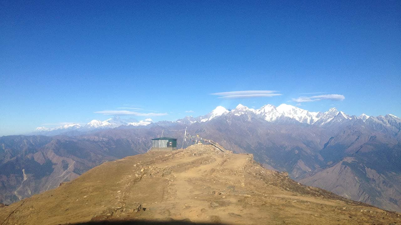 Scenic hilltop viewpoint with small shelter overlooking snow-capped mountain range under a clear blue sky.