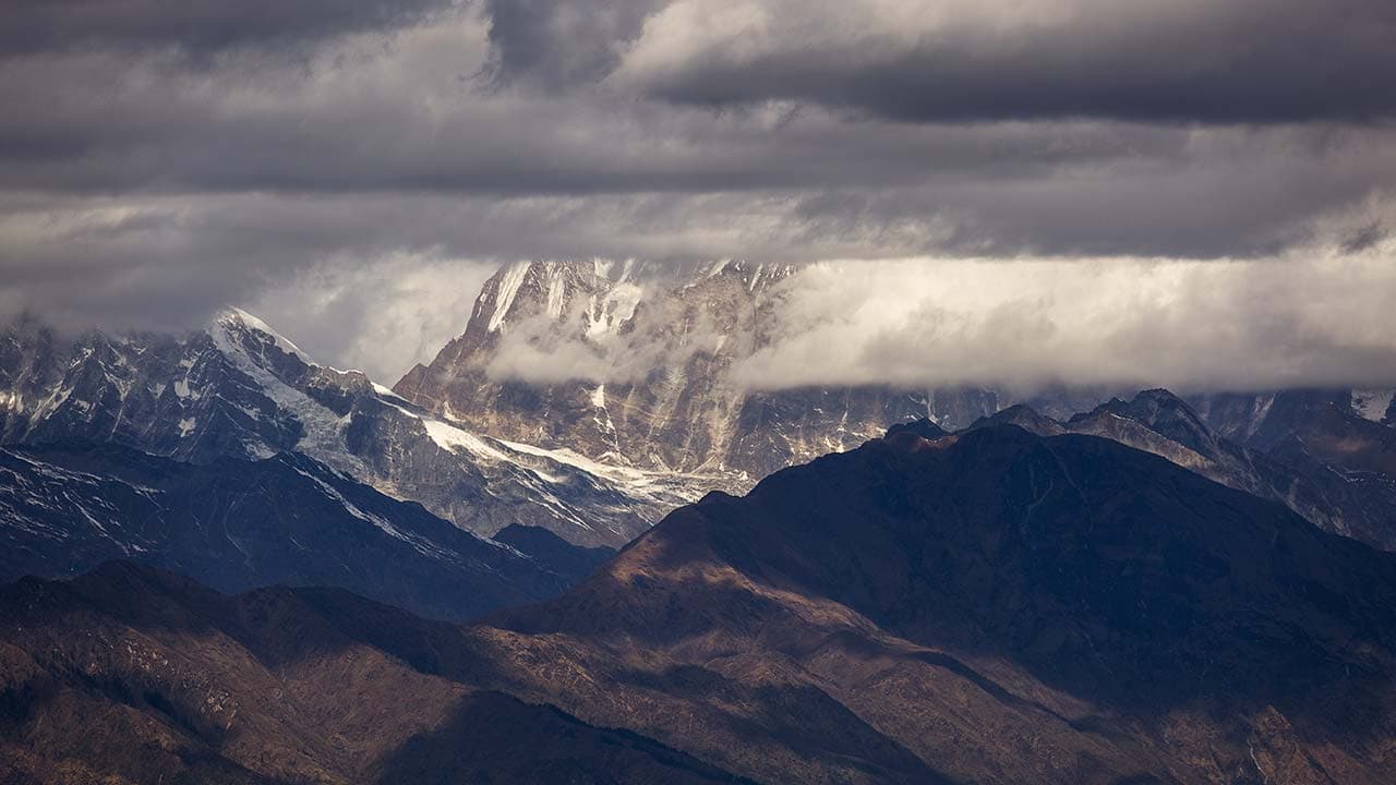 Panoramic view of dramatic snow-dusted mountain peaks emerging through dark cloud cover over deep Himalayan ridges.
