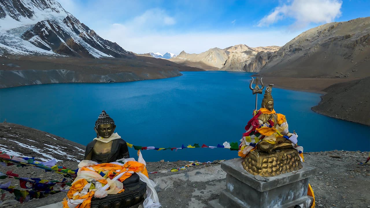 Buddha and Shiva statues with prayer flags overlooking turquoise Tilicho Lake in Nepal Himalayas.