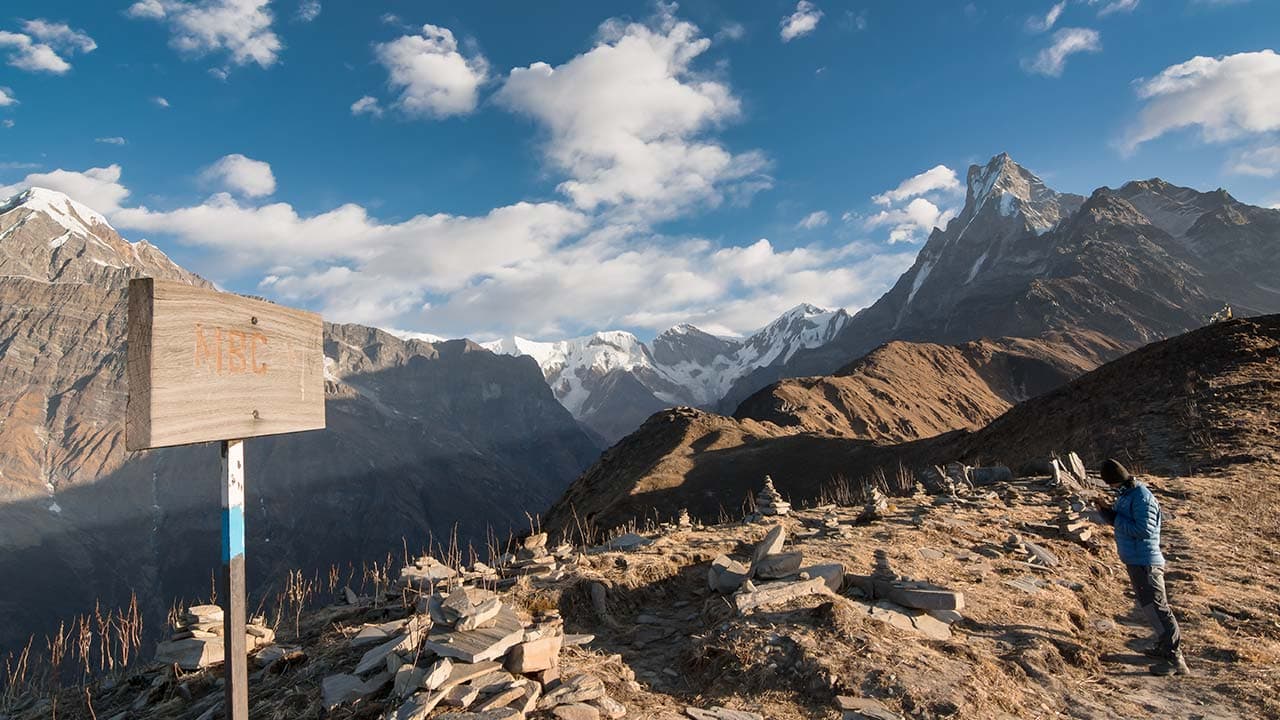 A wooden sign reading MBC on a mountain ridge with a trekker standing nearby and snow-capped peaks in the background.
