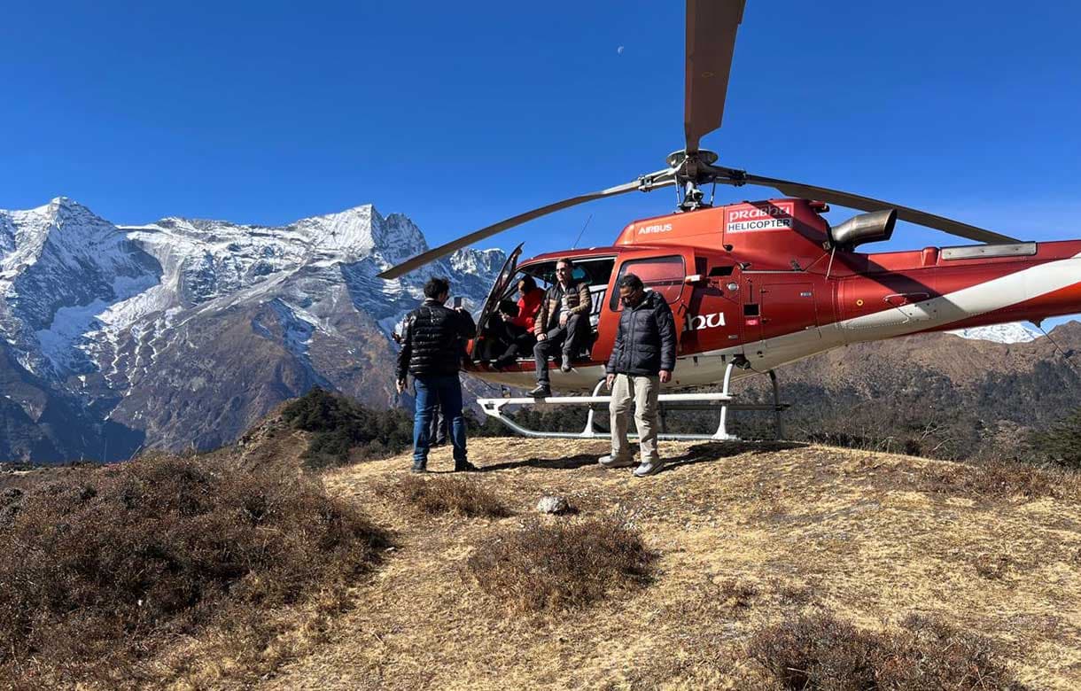 Red helicopter landed on a grassy ridge beside prayer flags and stone walls with Ama Dablam during an Everest Base Camp helicopter tour in Nepal.