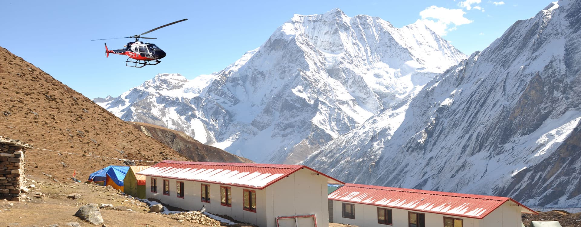 Red helicopter flying over mountain lodges with a snow-capped Himalayan peak in the background.