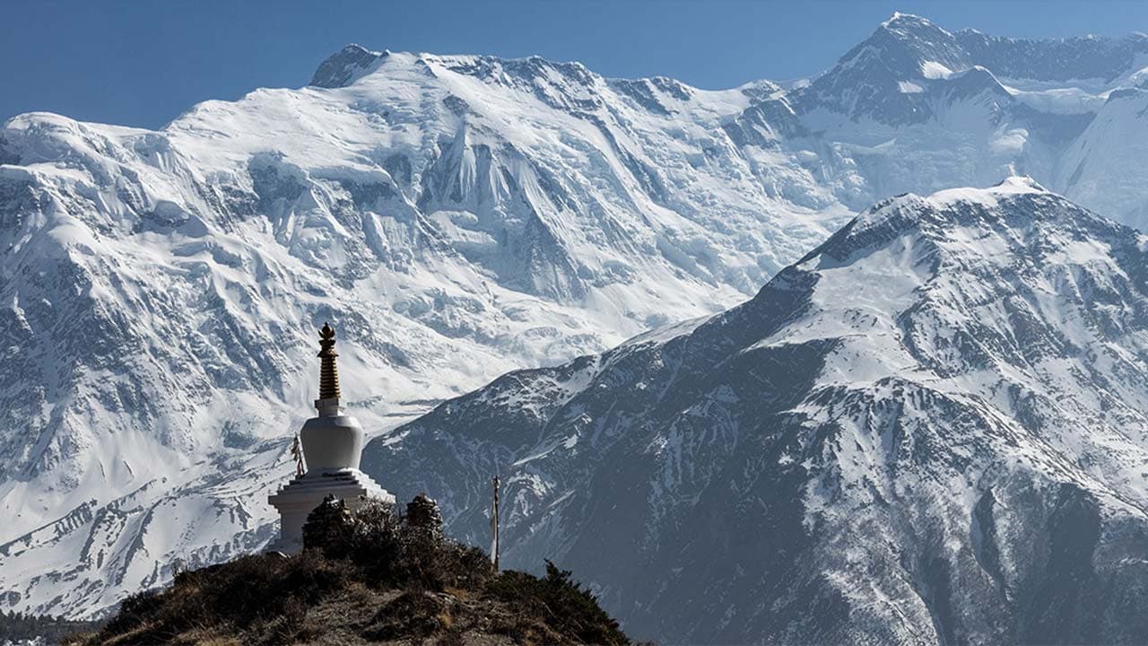 White Buddhist stupa standing on a hill against a backdrop of towering, snow-covered Annapurna mountain peaks under a clear blue sky.