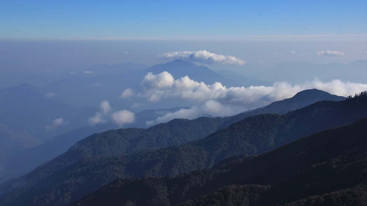 Panaromic view of rolling mountain ridges covered in clouds under a clear blue sky from the Mohare Danda Trek.
