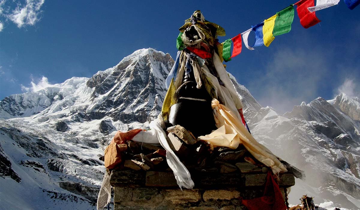 Stone chorten wrapped in colorful prayer flags with snow-capped Annapurna mountains in the background.