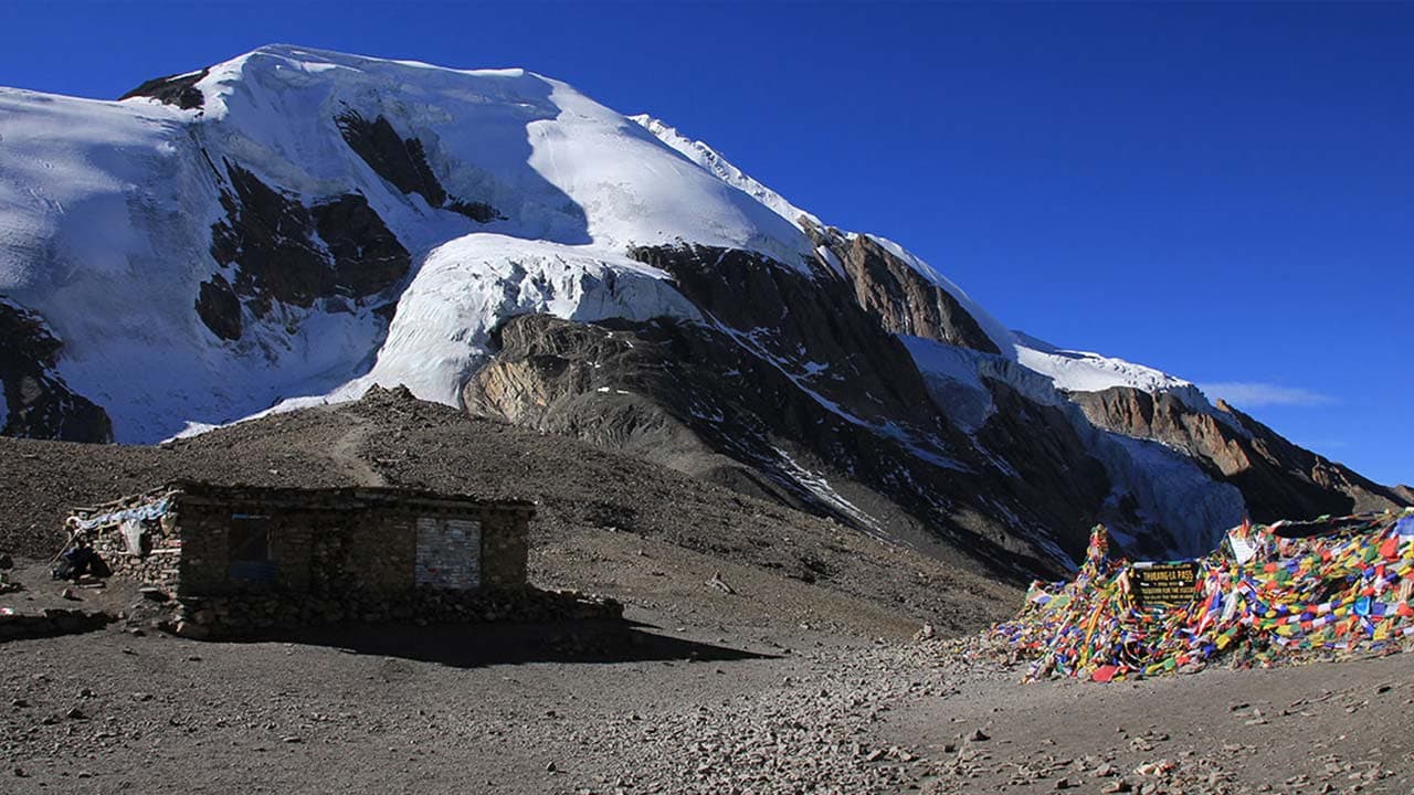 Small stone hut beside colourful prayer flags at Thorong La Pass with a snow-covered glacier peak under a clear blue sky.
