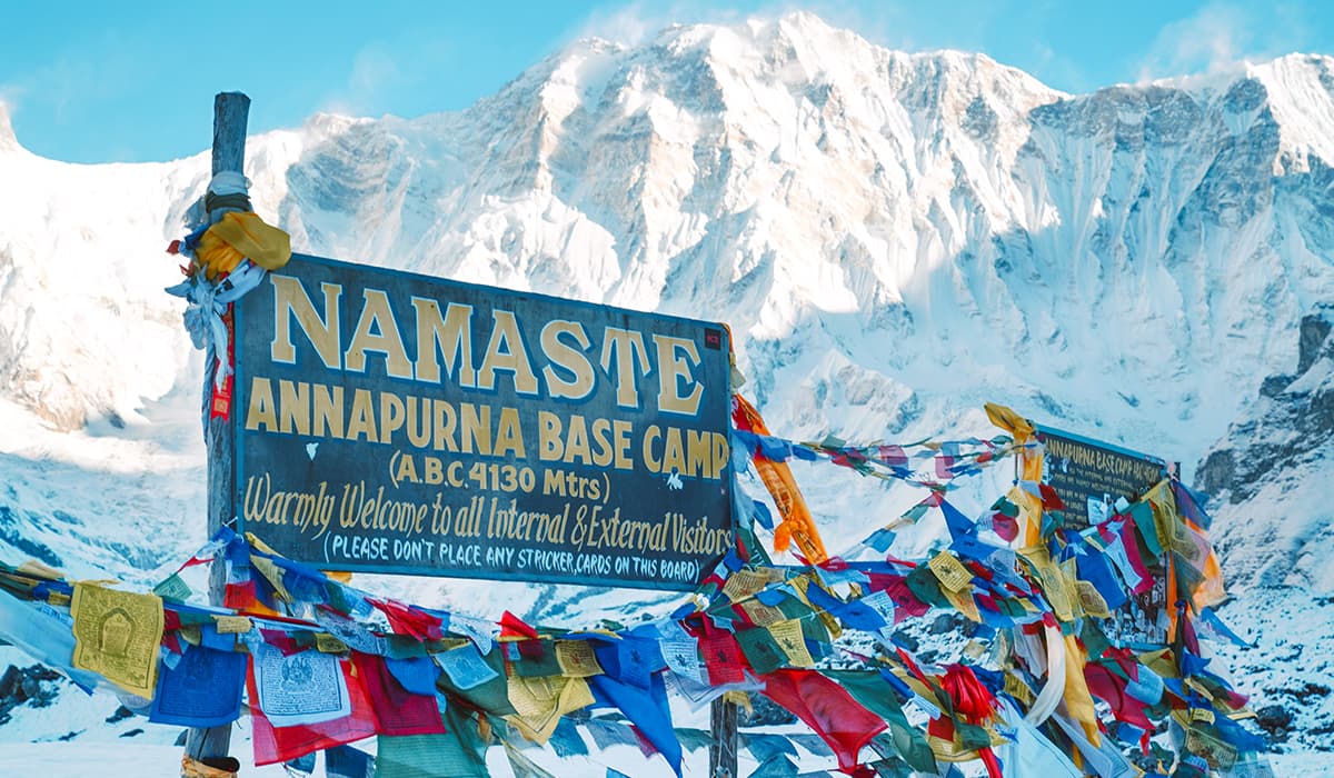 Wooden Annapurna Base Camp welcome signboard decorated with colorful prayer flags in front of snow-covered peaks.