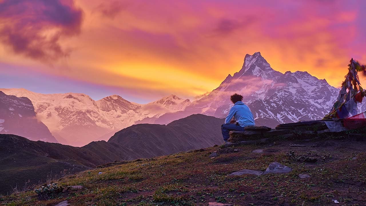 A trekker sits on a stone wall overlooking Mount Machhapuchhre under a colorful sunset sky.
