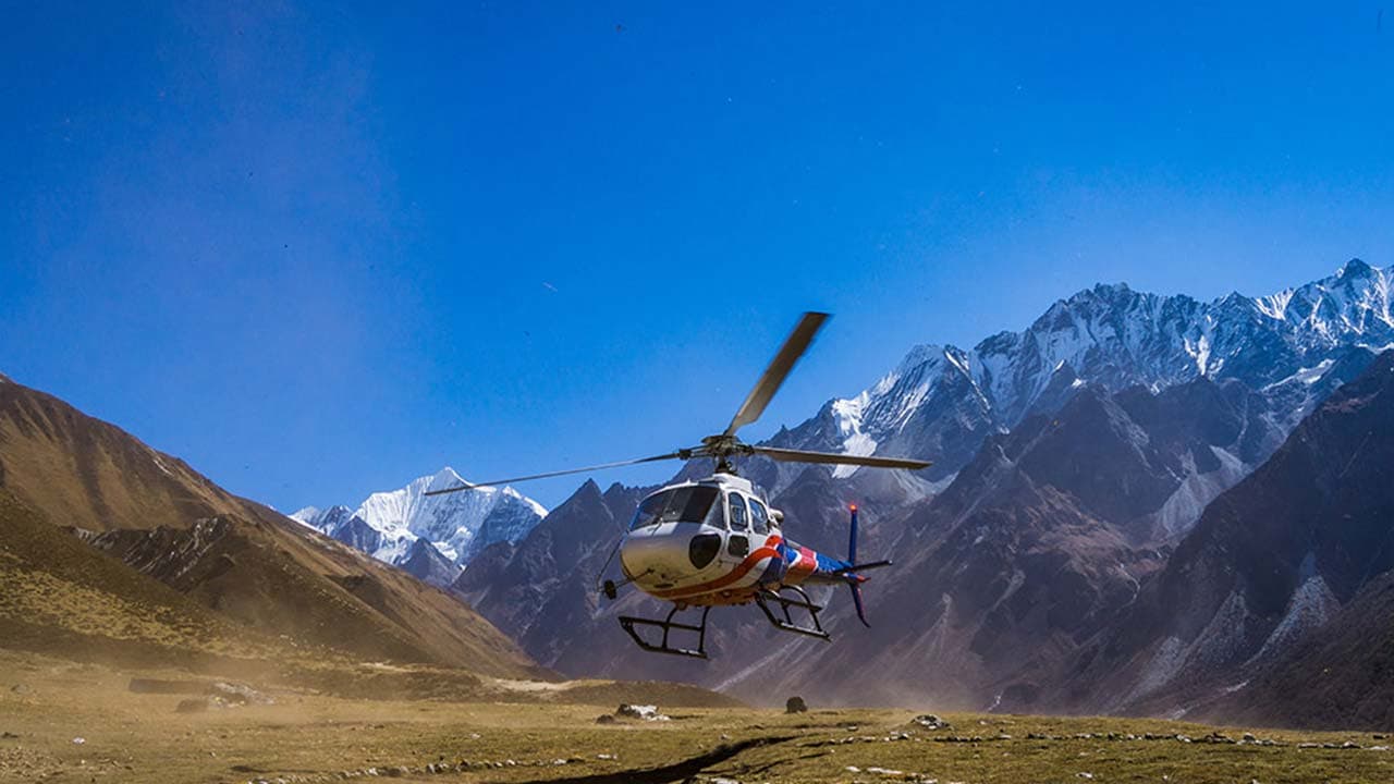 White and red helicopter hovering above a mountain valley with snow-capped Himalayan peaks behind it.