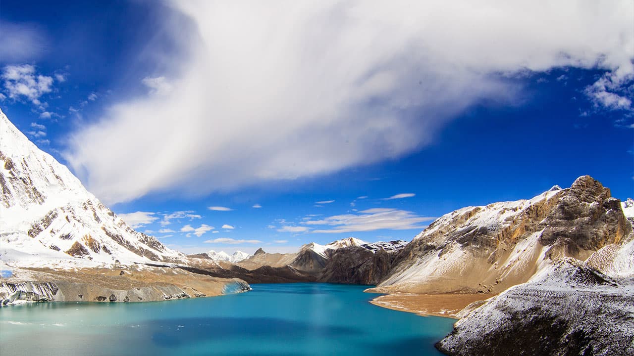 Turquoise Tilicho Lake surrounded by snow-capped mountains under a blue sky.
