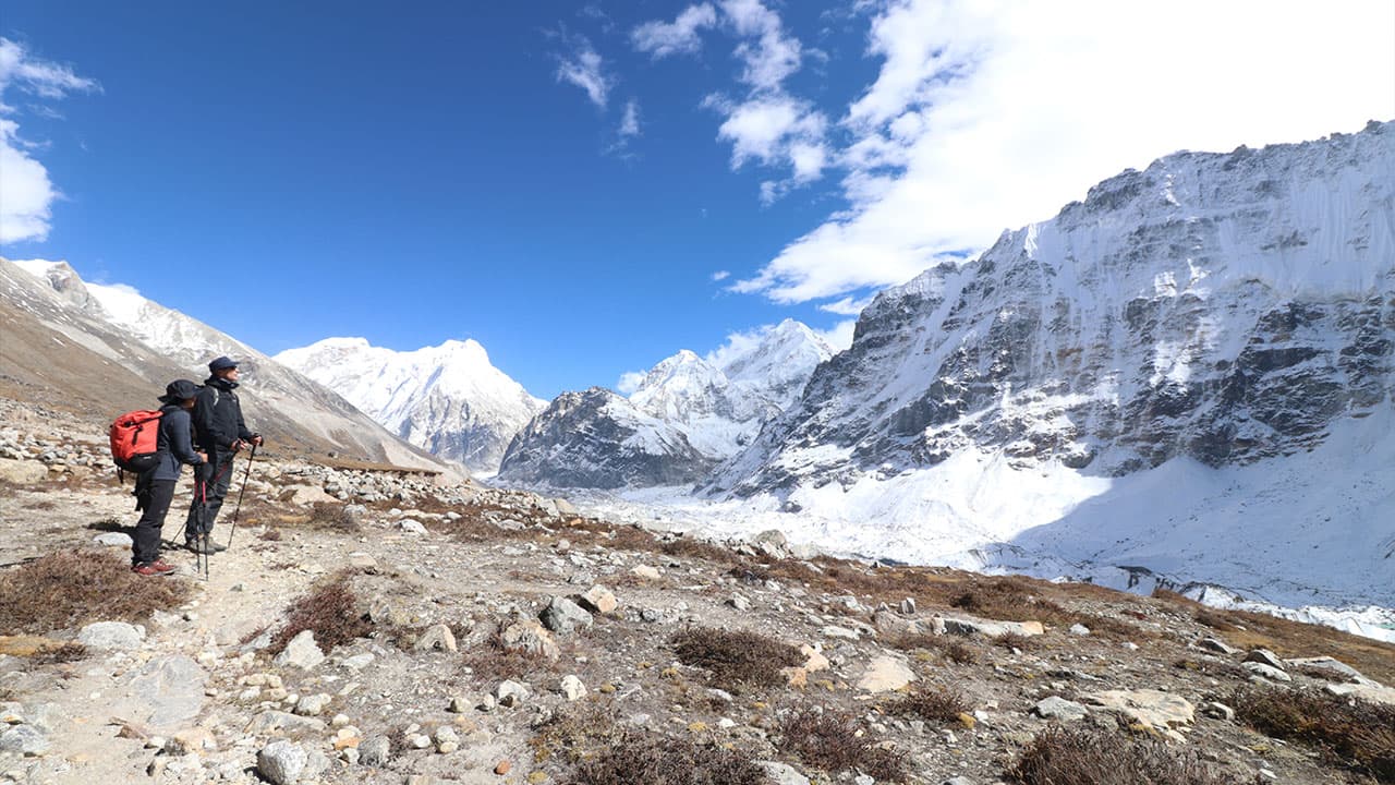 Two hikers on the Kanchenjunga Base Camp trekking trail with snowy mountains.