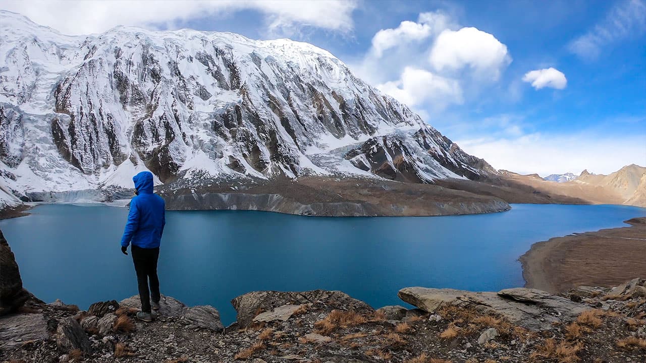 Trekker in blue jacket overlooking turquoise Tilicho Lake in Annapurna region Nepal under high mountain peaks.