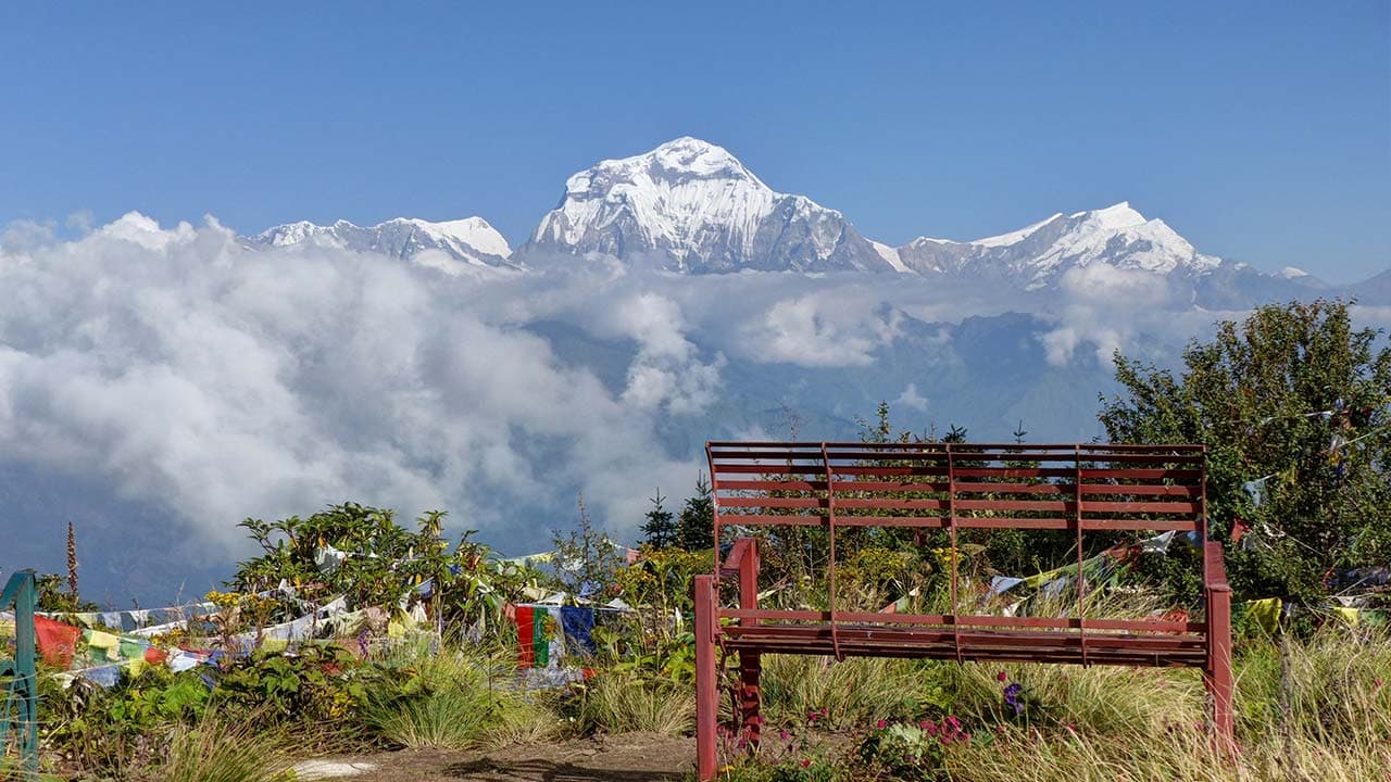Red wooden bench and prayer flags with snow-capped Dhaulagiri mountain over clouds under a blue sky.